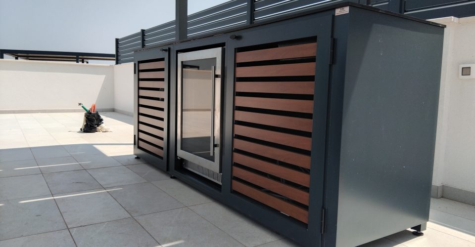 Vanjski hladnjak sa staklenim vratima ugrađen u tamnu aluminijsku kuhinju s drvenim panelima na otvorenoj terasi.
